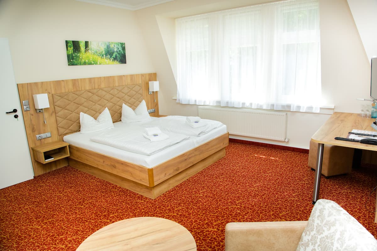 A hotel room with a wooden bed, a desk, a chair, a window with sheer curtains, and a painting on the wall.