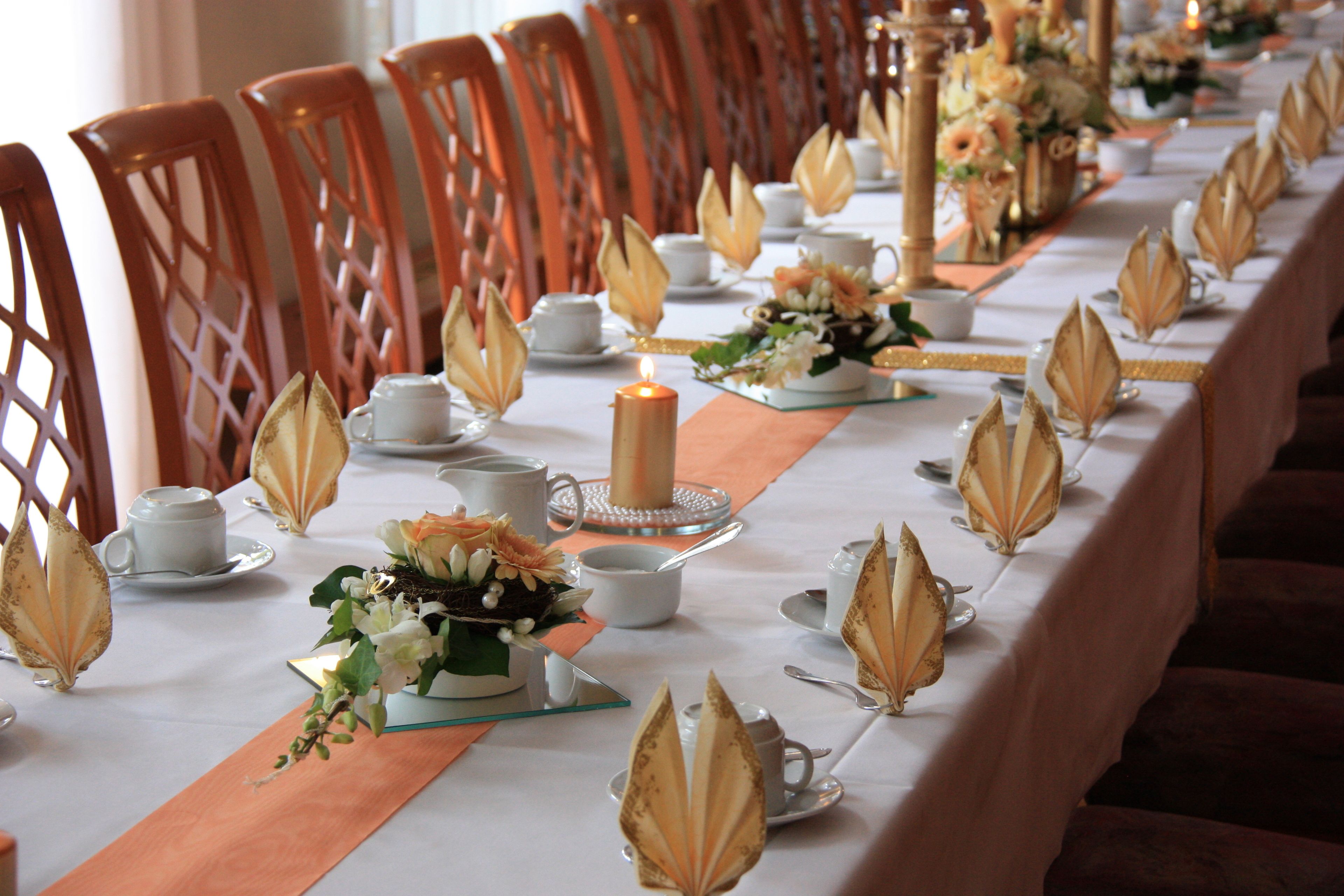 Una larga mesa de comedor decorada con mantel blanco, centros de mesa con pan de oro y servilletas naranjas.