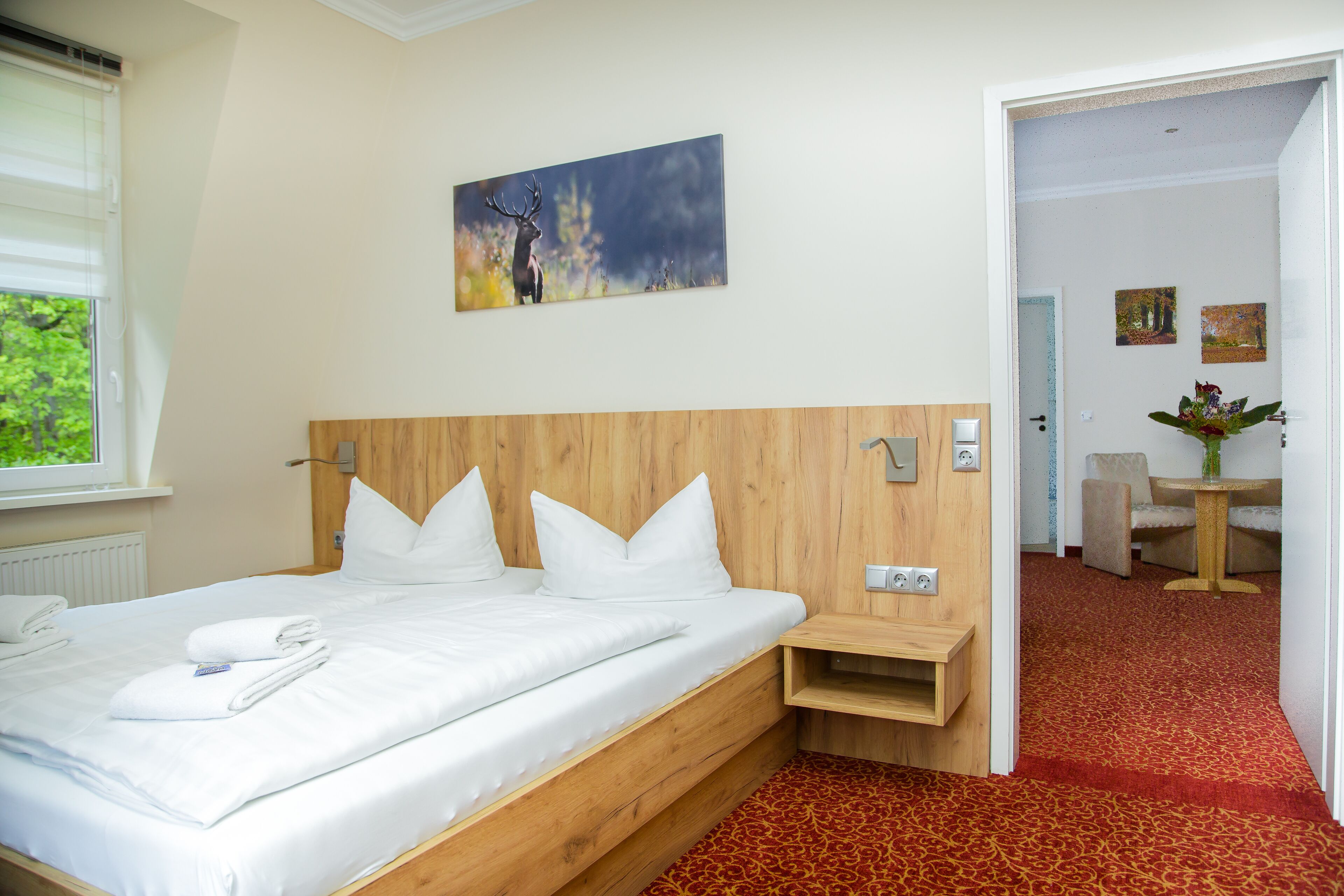 Habitación de hotel con cabecera de madera, ropa de cama blanca, un cuadro en la pared y vista al comedor.
