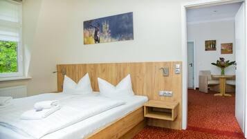 Habitación de hotel con cabecera de madera, ropa de cama blanca, un cuadro en la pared y vista al comedor.
