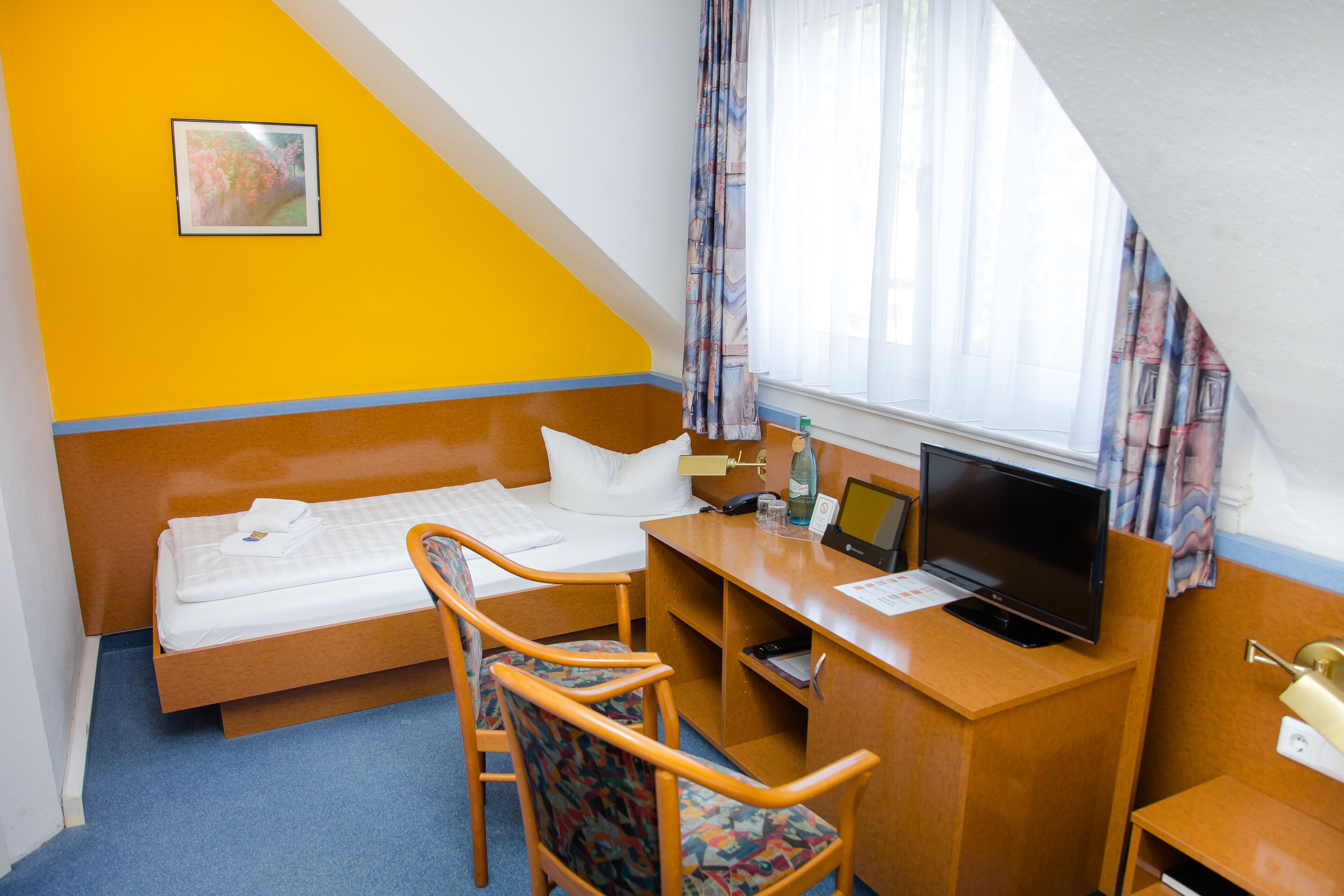 A hotel room with a bed, desk, chair, TV, and a painting on the wall.