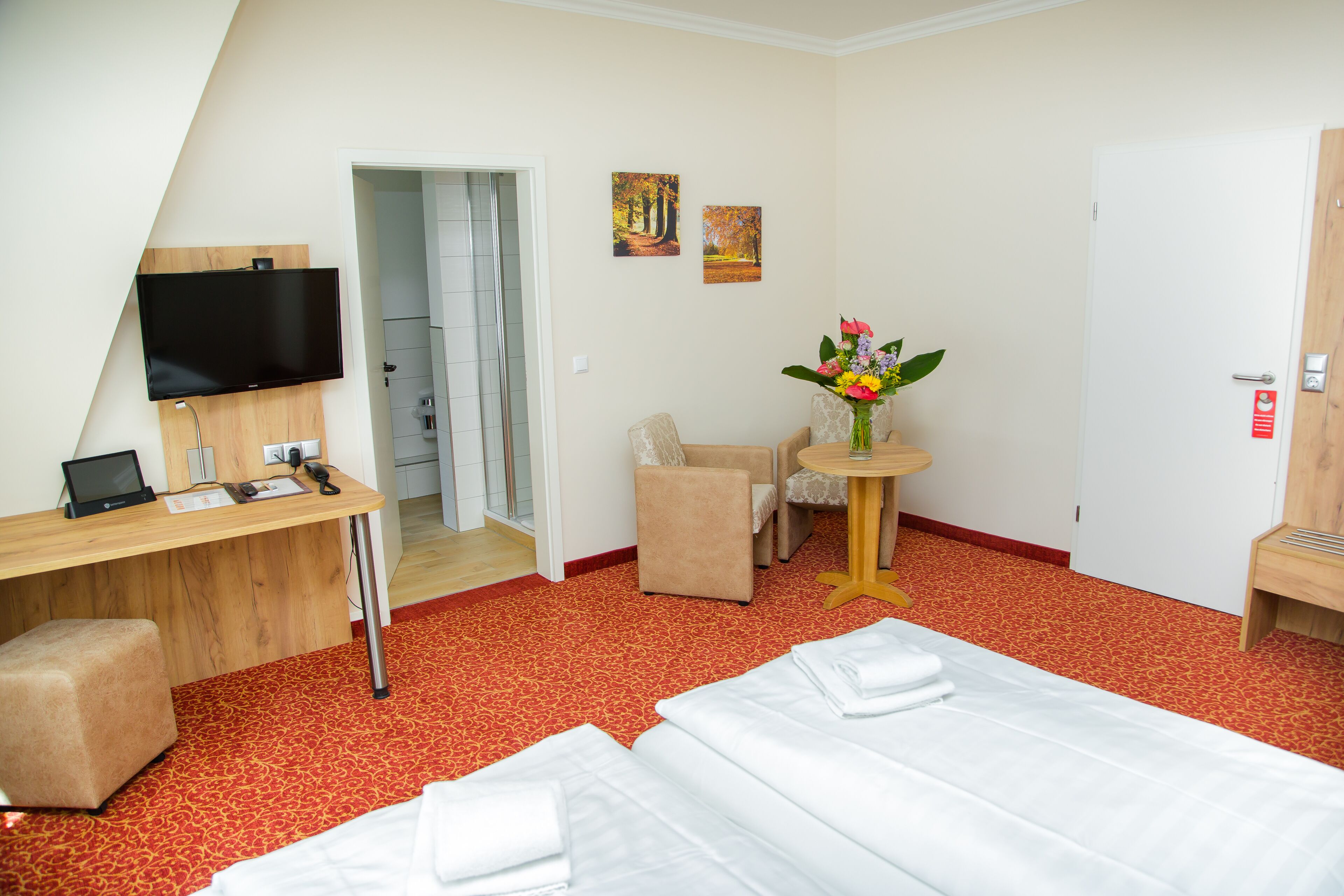 Habitación de hotel con cama, escritorio con televisor, silla, mesita con un florero y una puerta.