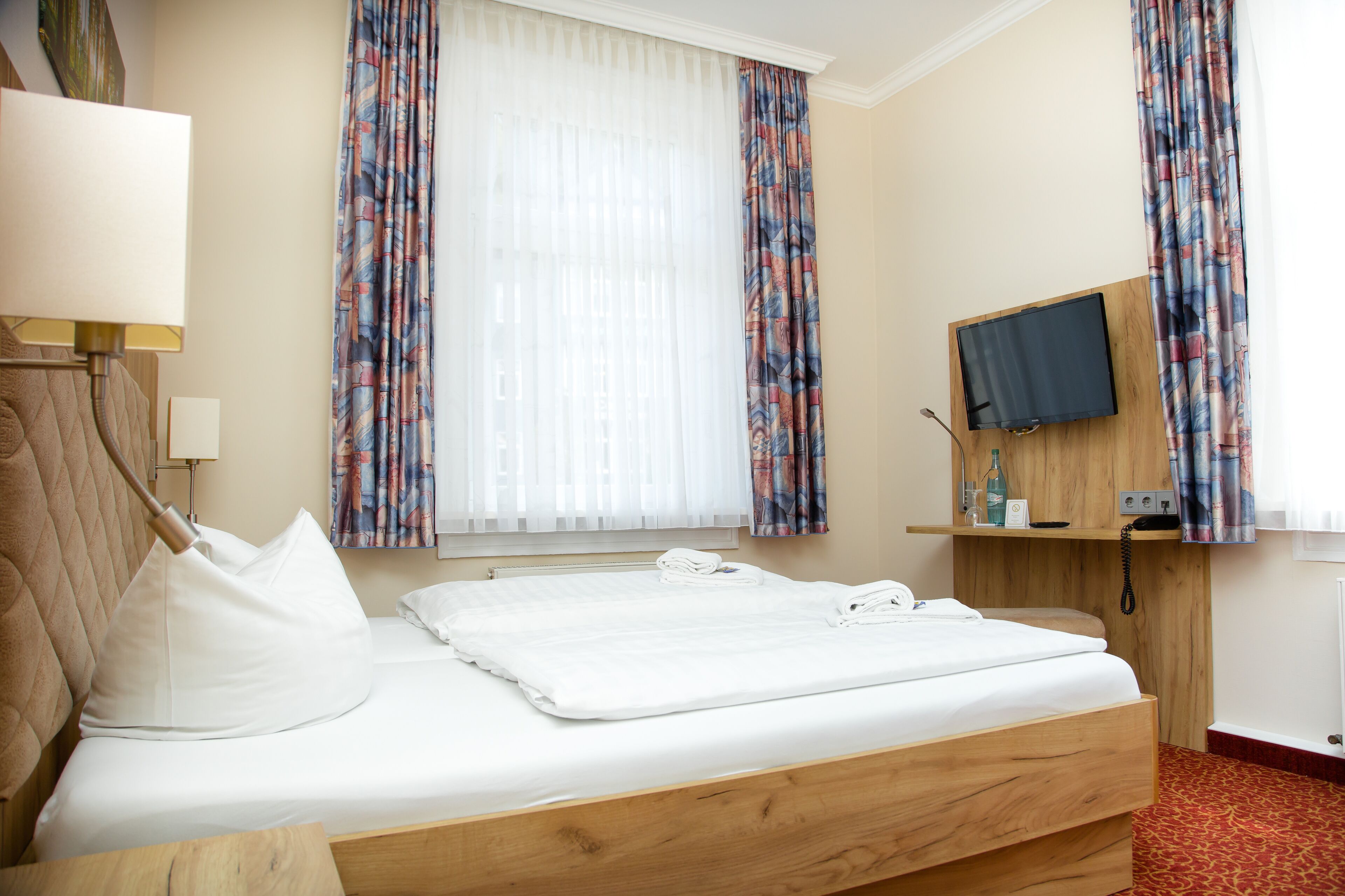 A hotel room with a bed, a TV mounted on the wall, a bedside table with a lamp, and curtains.