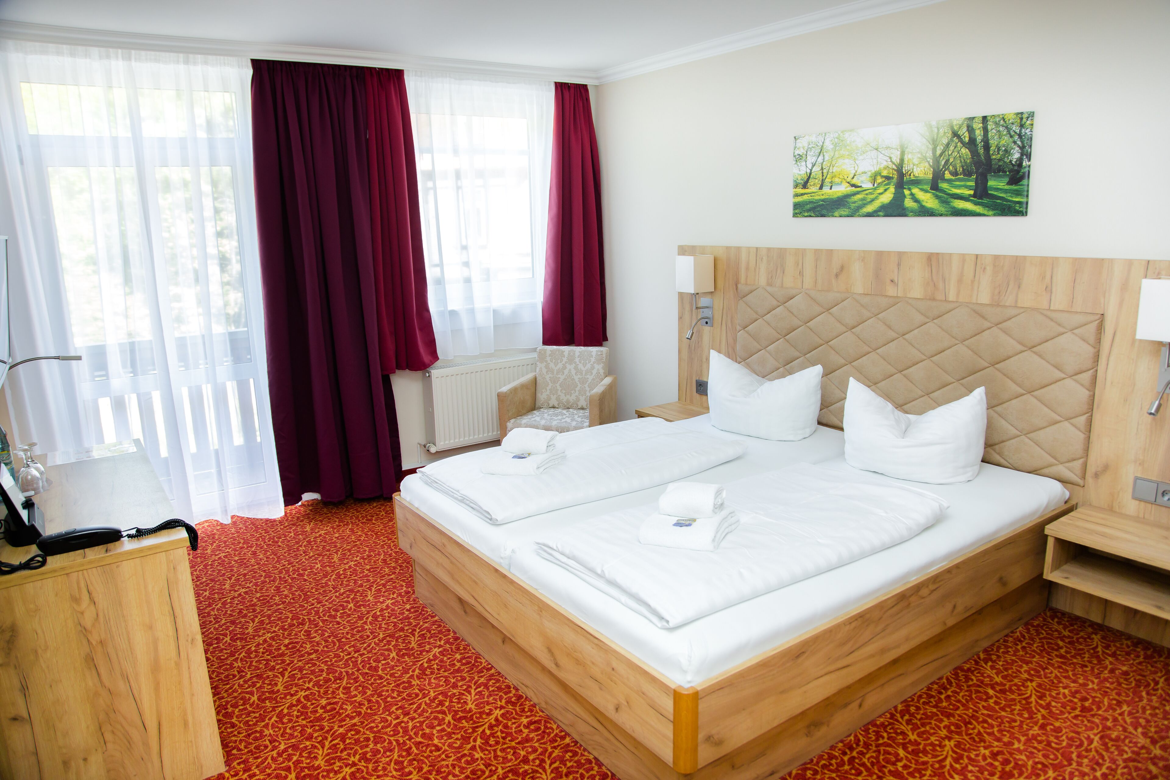 A hotel room with a large bed, two bedside tables, a wooden headboard, a radiator, a chair, and a window with curtains.
