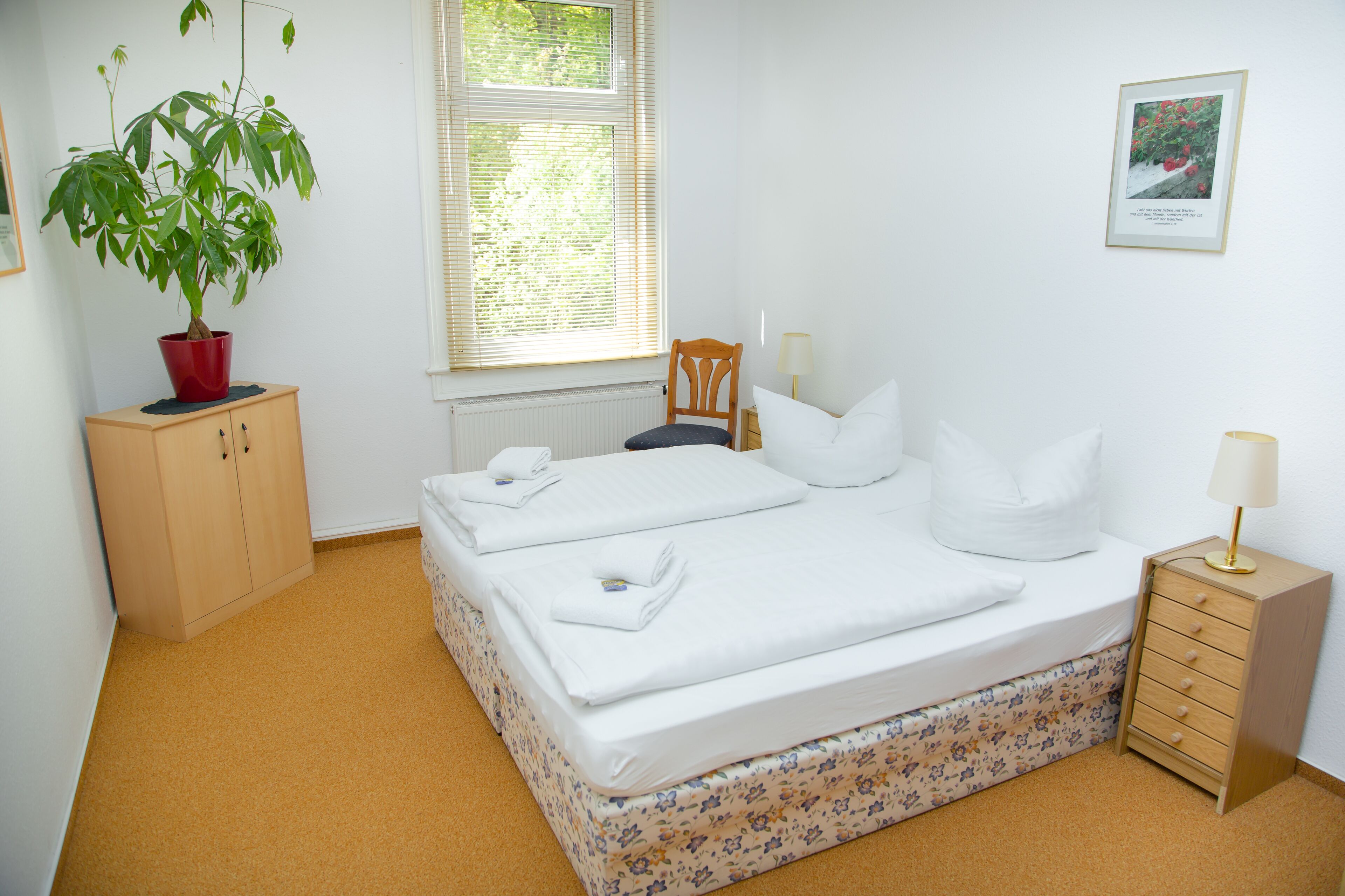 Une chambre à coucher avec un lit, une armoire en bois, une plante en pot, une chaise, une table de chevet avec une lampe et un tableau encadré au mur.