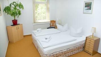 Ein Schlafzimmer mit einem Bett, einem Holzschrank, einem Blumentopf, einem Stuhl, einem Nachttisch mit Lampe und einem gerahmten Bild an der Wand.