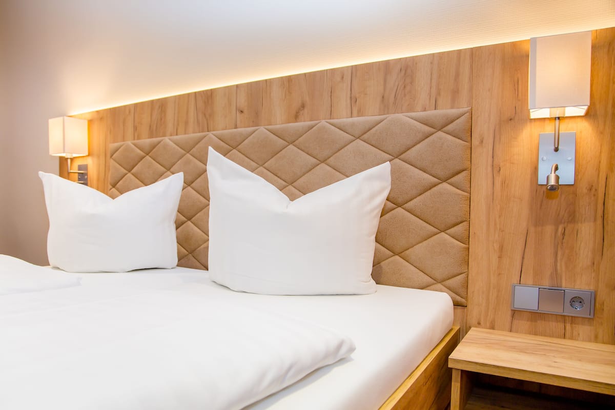 A neatly made bed with two pillows, a wooden headboard, bedside lamps, and a power outlet on the wall.