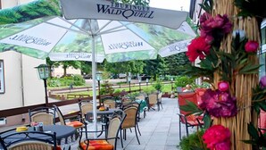 An outdoor patio with tables and chairs, covered by umbrellas with 'Thuringer WaldQuell' branding, and surrounded by greenery. - Waldhotel Friedrichroda (Friedrichroda)