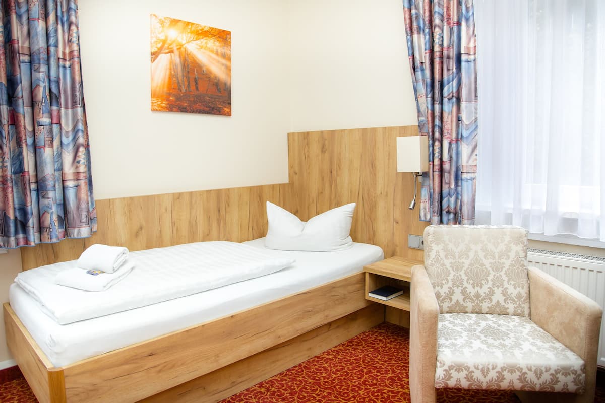 A single bed with a wooden headboard, a bedside table with a lamp, a patterned armchair, and a painting on the wall.