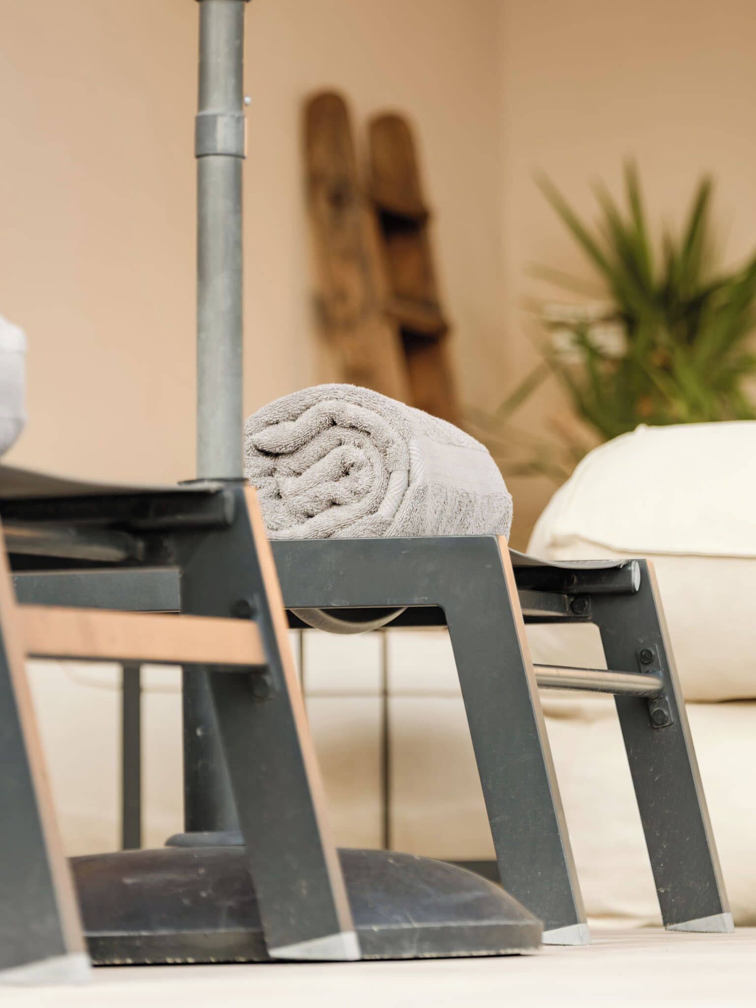Una toalla sobre un soporte metálico, una silla de madera y un cojín blanco en una habitación con pared color beige y una planta.