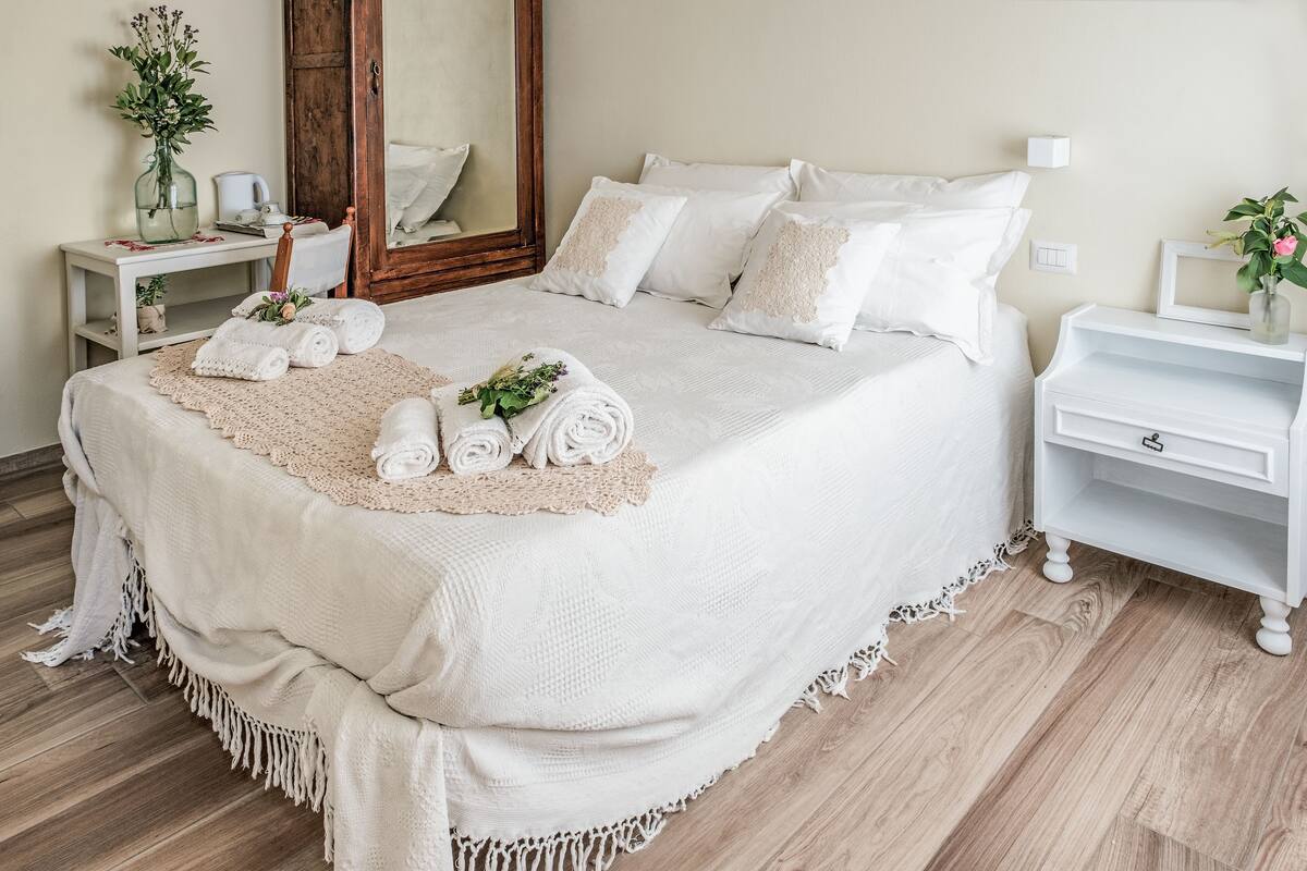 Ein ordentlich bezogenes Bett mit weißer Bettwäsche und einer Spitzendecke, ein Nachttisch mit Blumen in einer Vase sowie ein Spiegel über dem Bett.