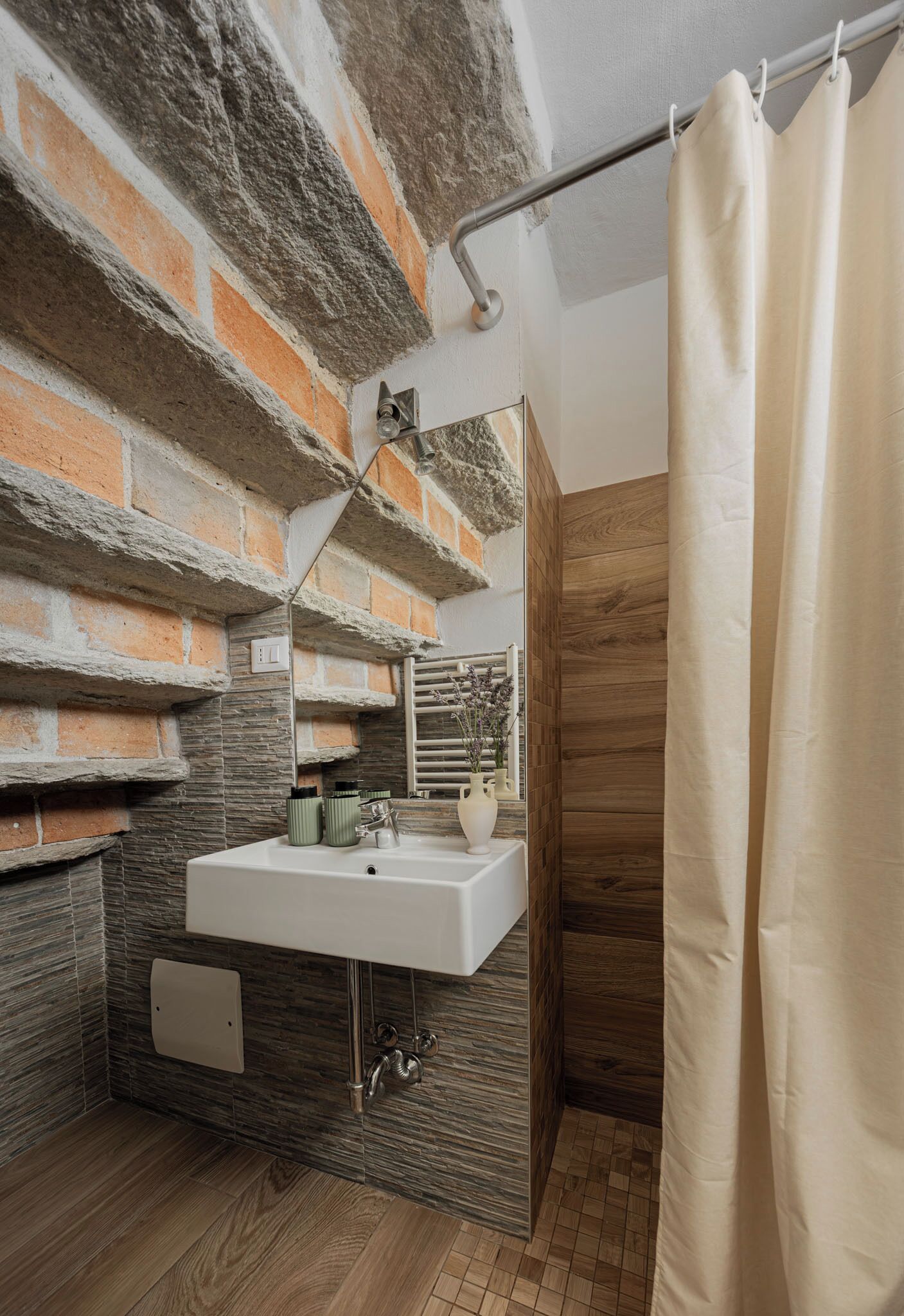 Un bagno con parete in pietra, pavimento in legno, lavandino bianco e tenda per la doccia.