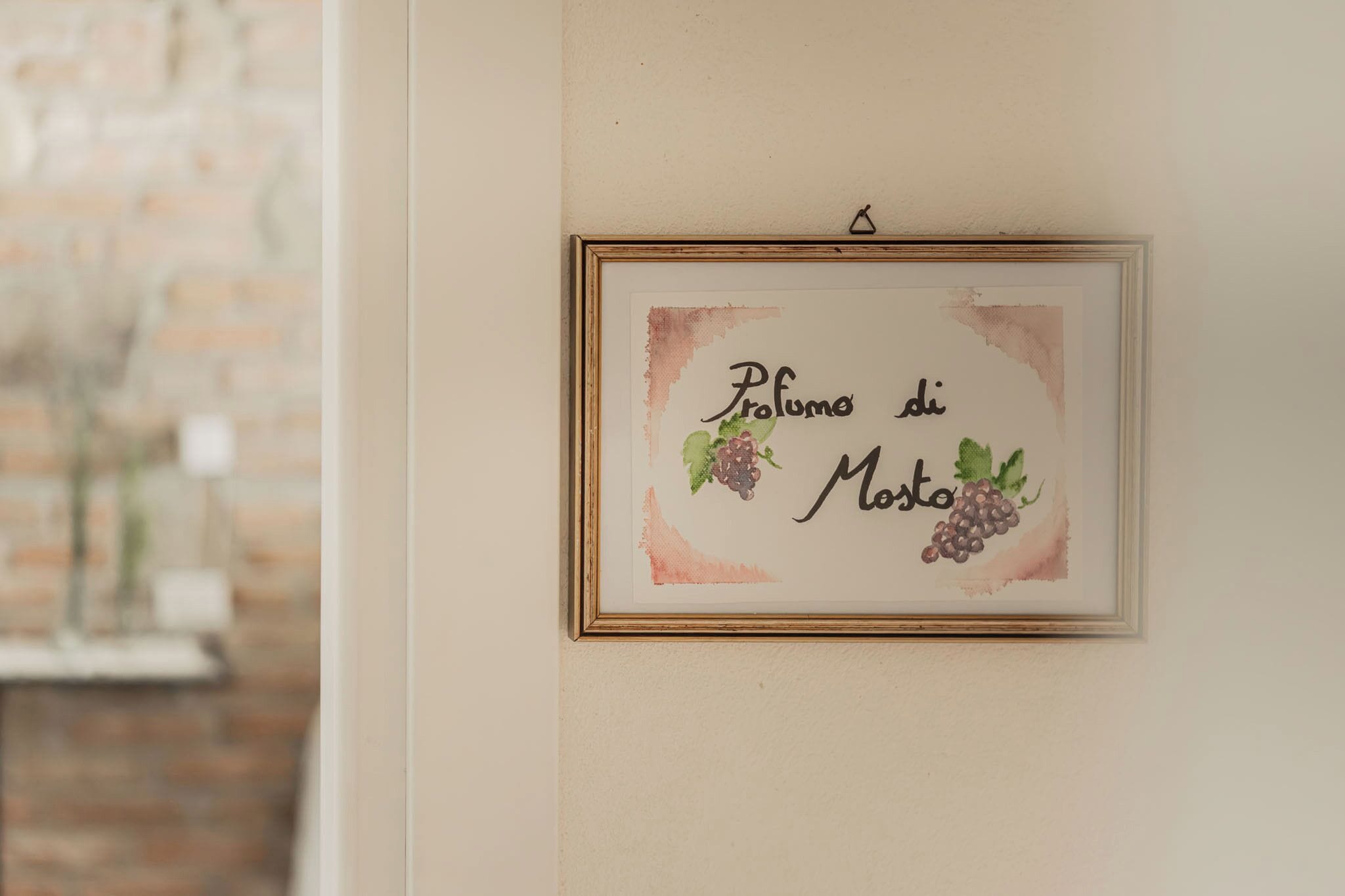 Obra emoldurada com o texto 'Profumo di Mosto' e ilustrações de uvas em uma parede.
