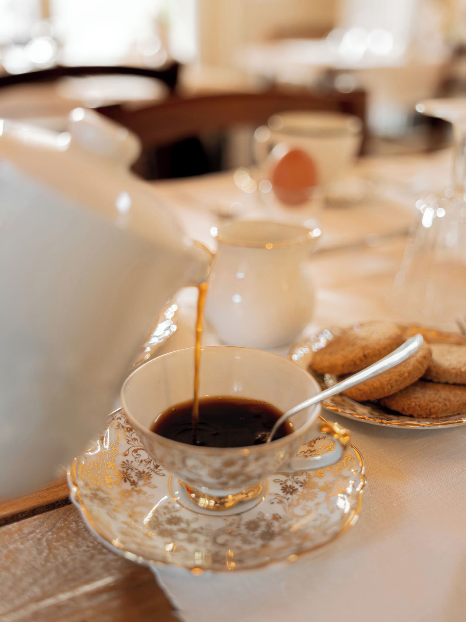 Una taza de café siendo servida en un platito, un plato con galletas y una jarrón al fondo.