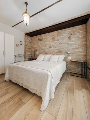 Un dormitorio con pared de ladrillo, suelo de madera, cama con ropa blanca de cama y una lámpara colgante.