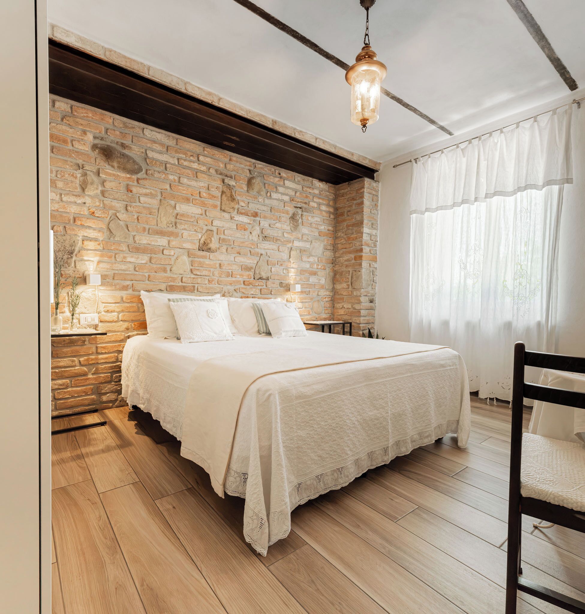 A bedroom with a brick wall, a wooden floor, a bed with white bedding, a hanging light fixture, and a window with sheer curtains.
