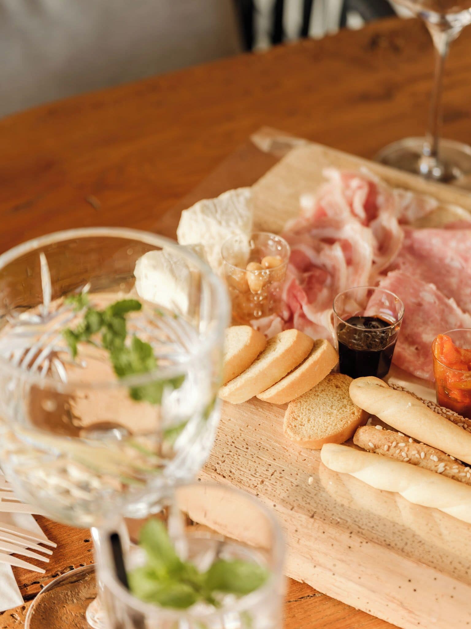 Ein Holztisch mit einer Wurst- und Käseplatte, bestehend aus verschiedenen Fleischsorten, Käsesorten und Brot, begleitet von Gläsern Wasser und einem dunklen Getränk.