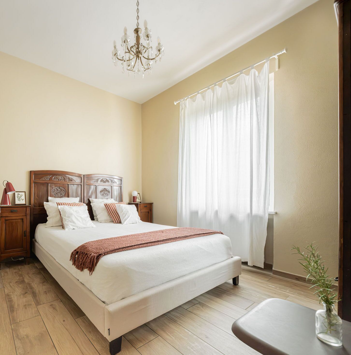 Une chambre avec un grand lit, une tête de lit en bois, du linge de lit blanc, un lustre, une fenêtre avec des rideaux et une table de chevet avec une lampe.