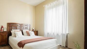Une chambre avec un grand lit, une tête de lit en bois, du linge de lit blanc, un lustre, une fenêtre avec des rideaux et une table de chevet avec une lampe.