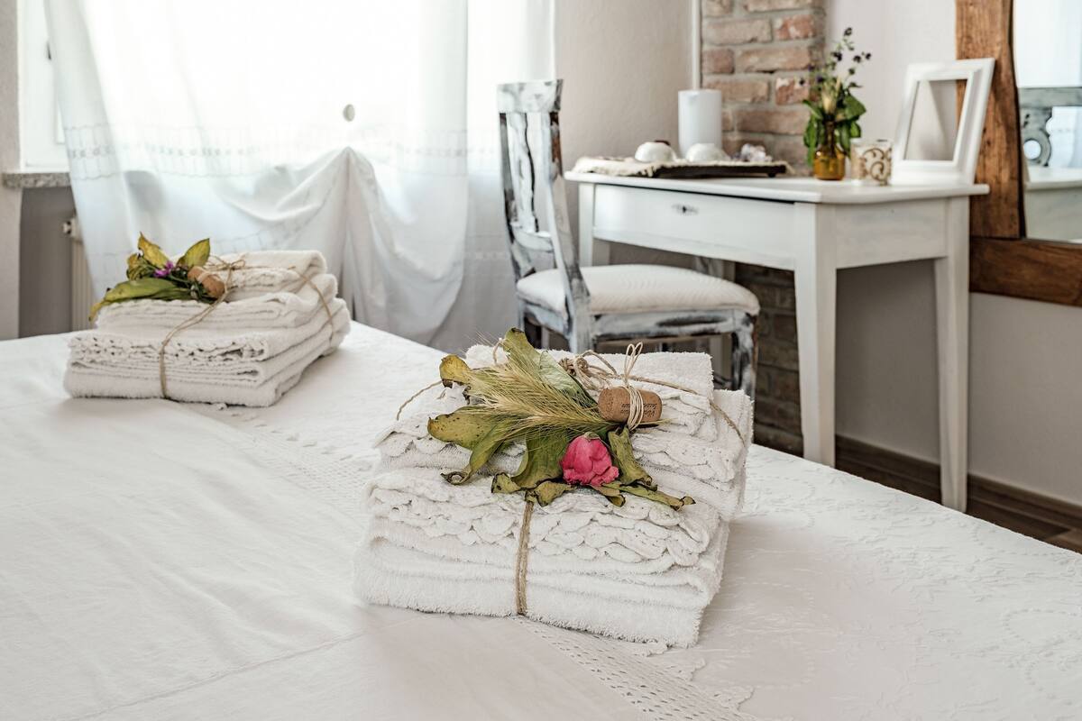 Ein ordentlich bezogenes Bett mit gefalteten Handtüchern und dekorativen Blumen, ein weißer Stuhl und ein kleiner Tisch mit einer Vase und Bilderrahmen in einem Zimmer mit Backsteinwand und weißen Vorhängen.