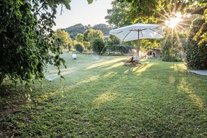 Un jardín trasero con césped bien cuidado, con una silla de patio y una sombrilla, rodeado de árboles y vistas a colinas al fondo.