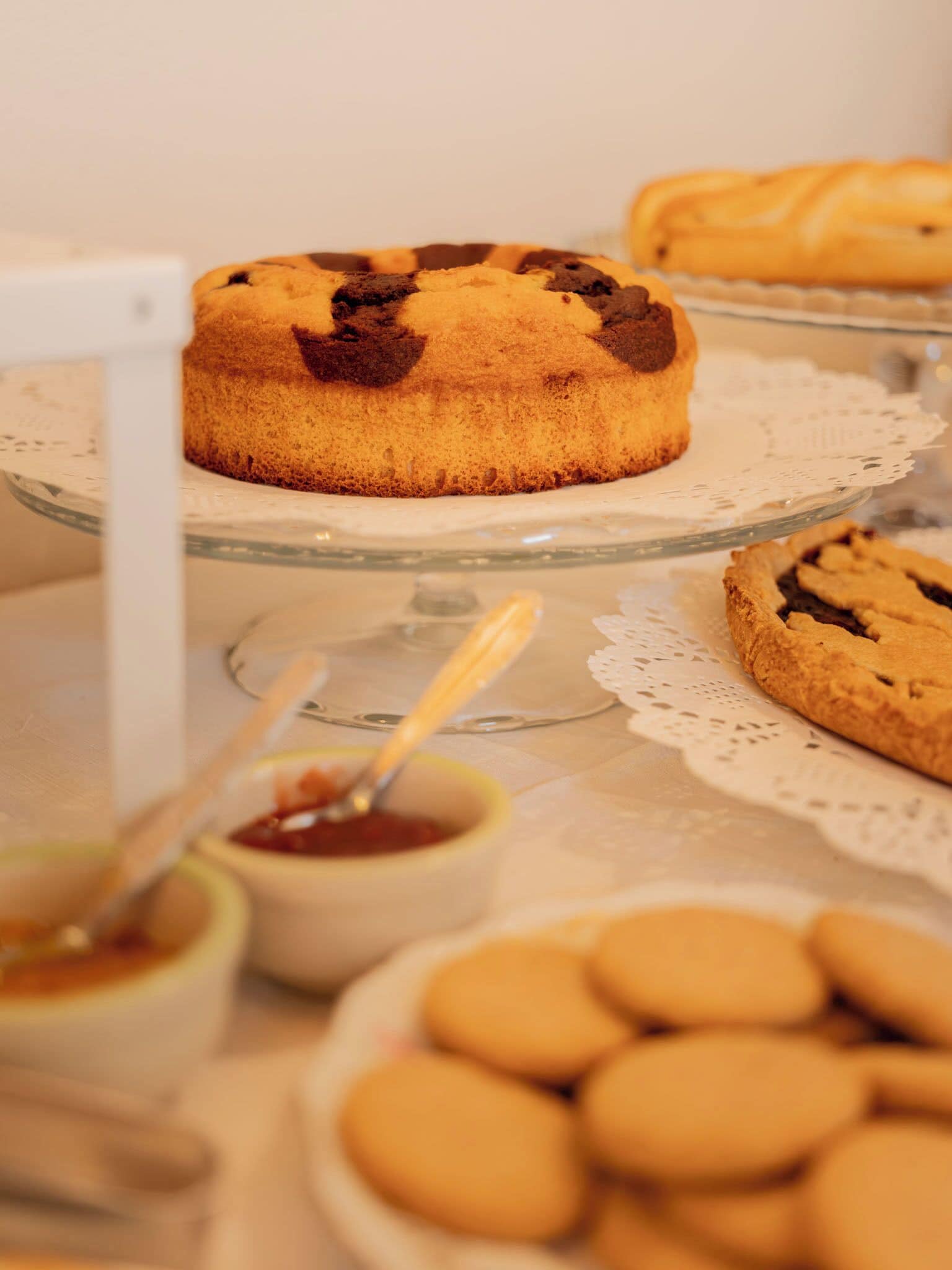 A dessert table with a chocolate chip muffin on a glass plate, a jar of jam, and several cookies.