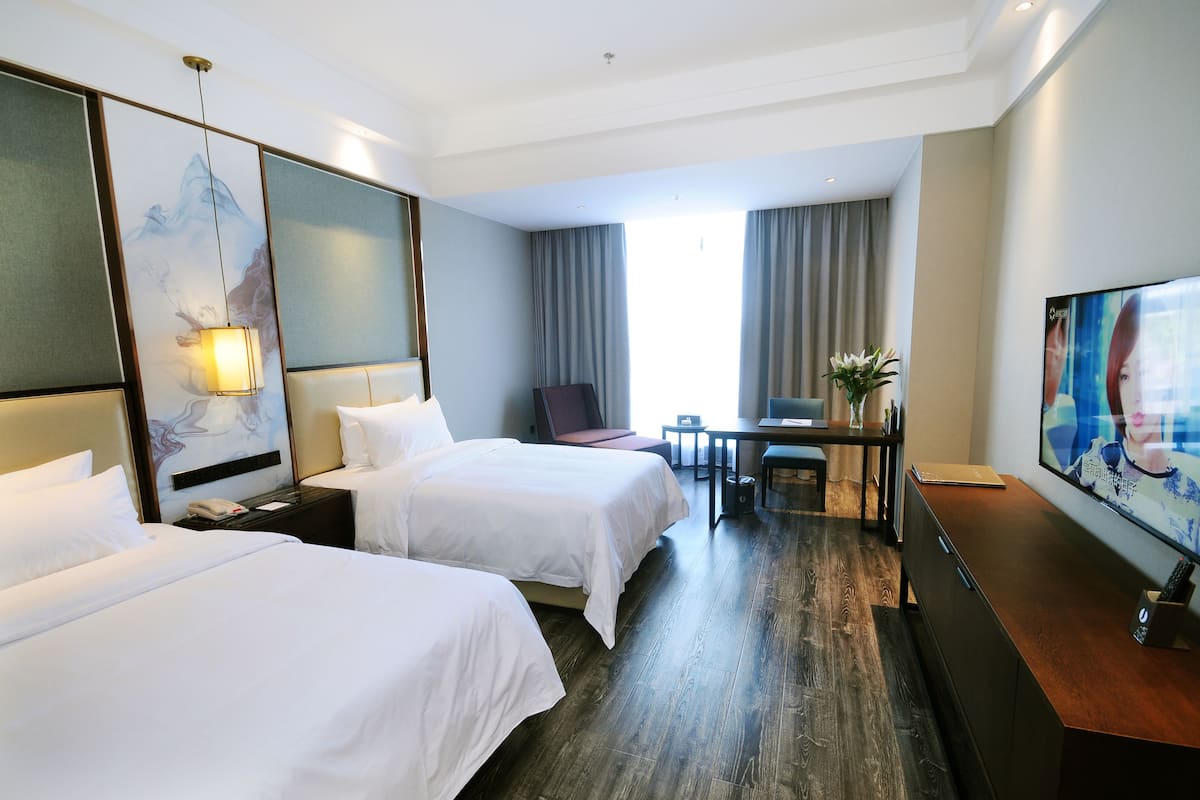 Ein Hotelzimmer mit zwei Betten, einem Schreibtisch, einem Stuhl, einem Fernseher und einem großen Fenster mit Vorhängen.