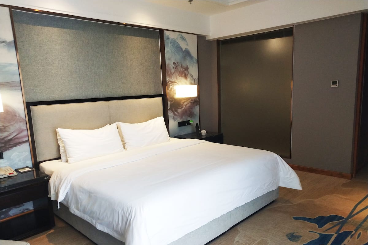Ein Hotelzimmer mit einem großen Bett, einem Nachttisch mit Telefon und Lampe, einem Bild an der Wand und einem Schrank.