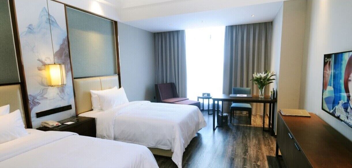 A hotel room with two beds, a desk, a chair, a lamp, and a painting on the wall.