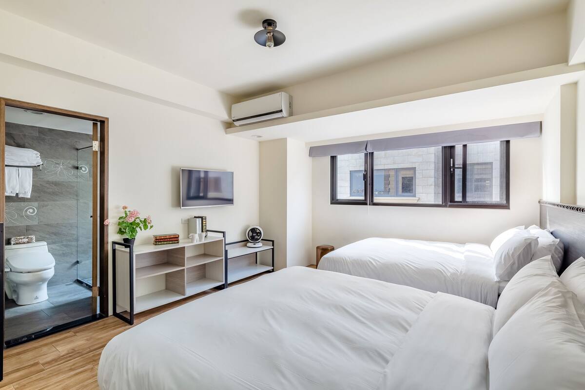 A modern hotel room with two beds, a TV, a small shelf with books and a vase, and a window with a view of the city.