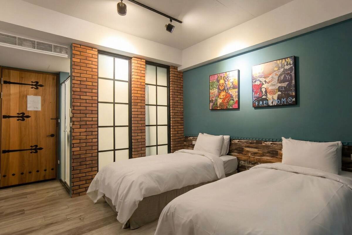 A room with two beds, a wooden door, a brick wall, and two artworks on the wall.
