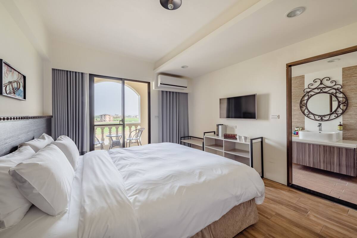 A hotel room with a large bed, a TV, a bathroom with a mirror, and a balcony with outdoor furniture.