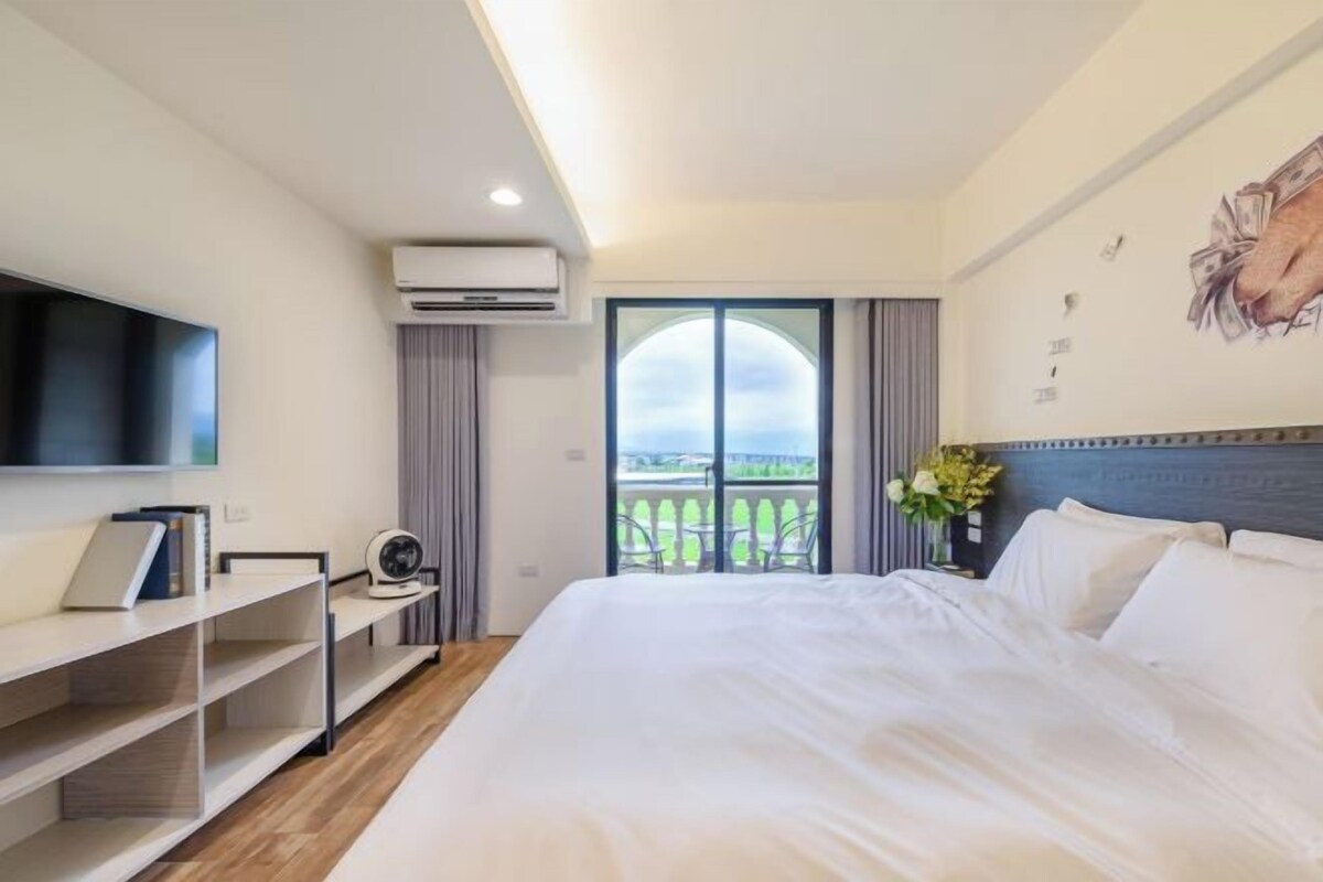 Habitación de hotel con cama, televisor de pantalla plana, una mesita con un ventilador y balcón con vistas.