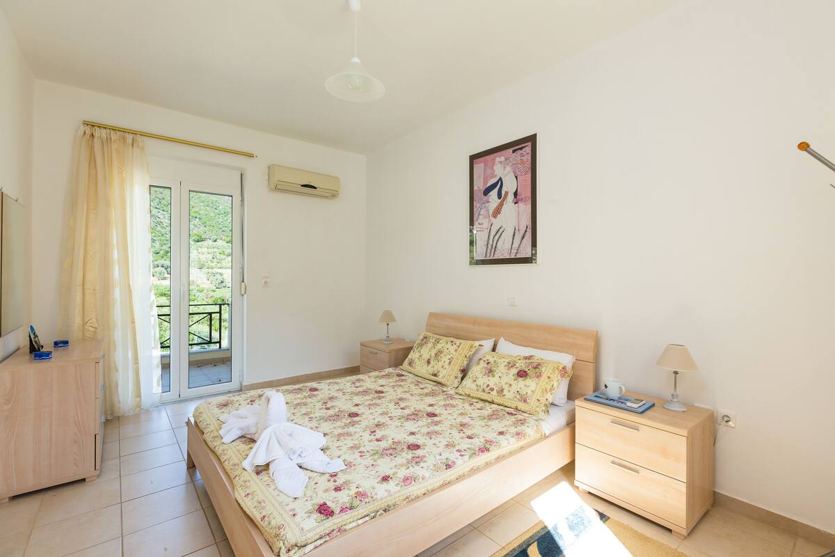Quarto com cama, mesas de cabeceira, penteadeira, janela com cortinas e um quadro na parede.