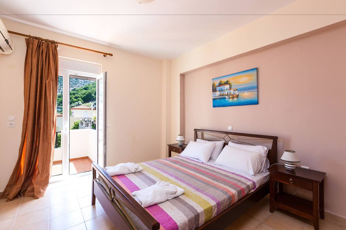 Quarto com cama, mesa de cabeceira, quadro e vista para o exterior.