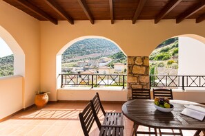 Une terrasse avec une table en bois, des chaises et un saladier rempli de fruits. Deux fenêtres en arc offrent une vue sur une colline et des bâtiments.