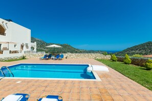 A swimming pool with lounge chairs and umbrellas, surrounded by a stone wall and greenery, with a view of the sea and hills.