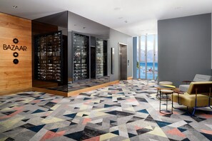 A modern lobby with a geometric patterned floor, a wooden wall with 'BAZAAR' signage, a wine display, and a seating area with a view of the sea.