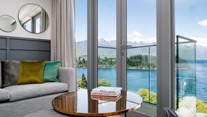 A modern living room with a grey sofa, a round wooden coffee table, and a view of a lake and mountains through large glass doors. - QT Queenstown (Queenstown)