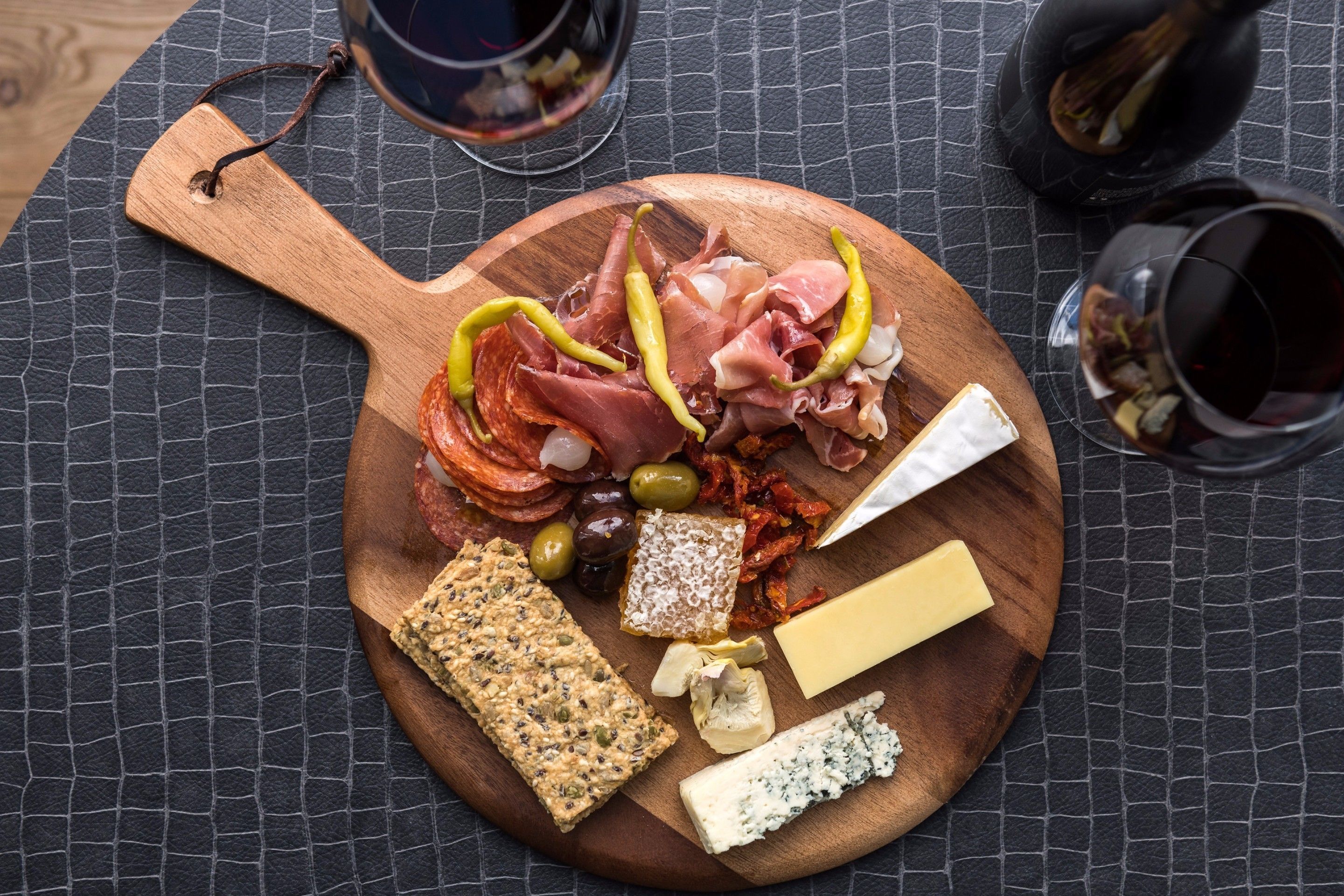 A wooden board with various cheeses, cured meats, olives, and crackers on a textured tablecloth.