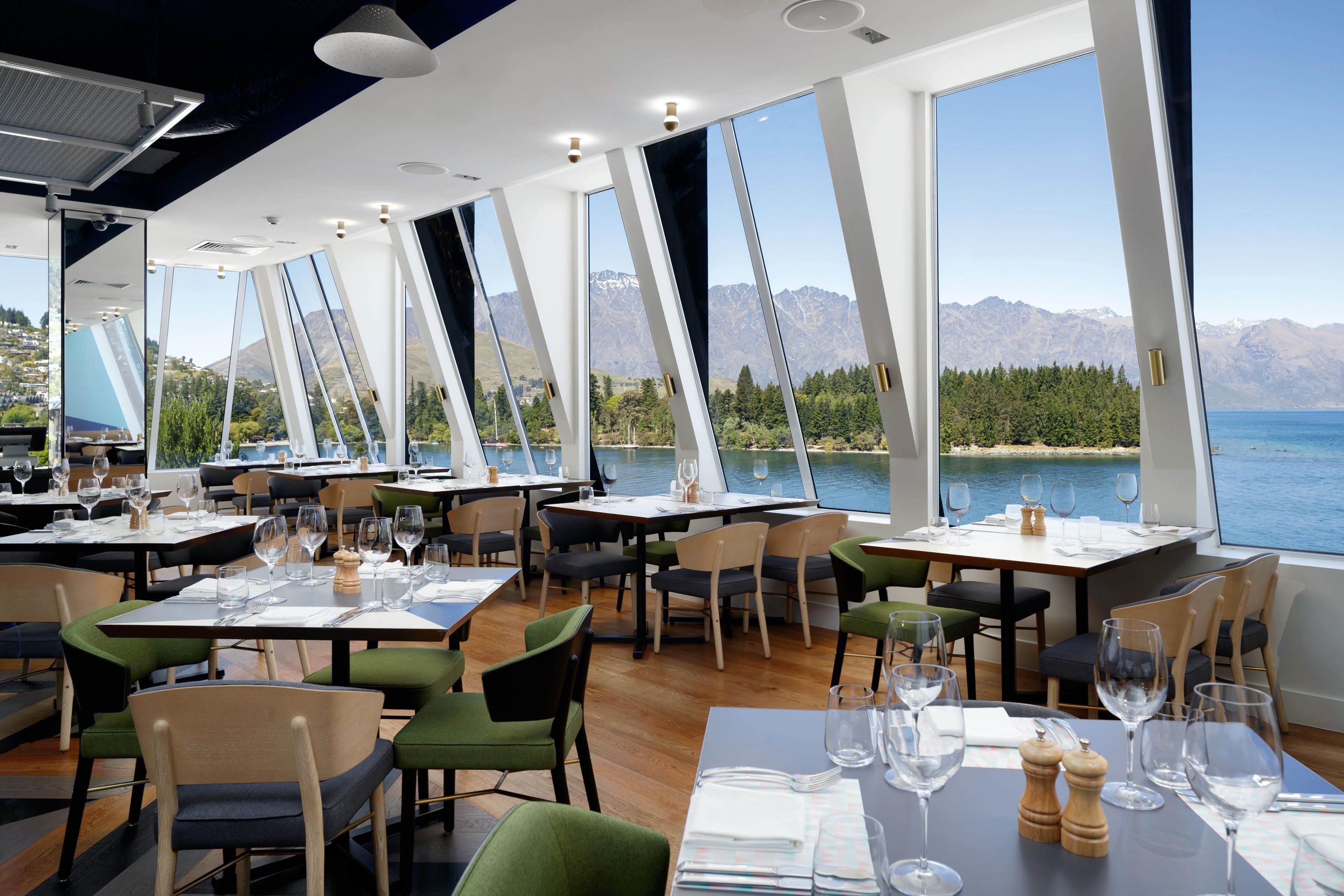 Un restaurante con mesas dispuestas para comer, amplias ventanas que ofrecen vistas a montañas y un lago.
