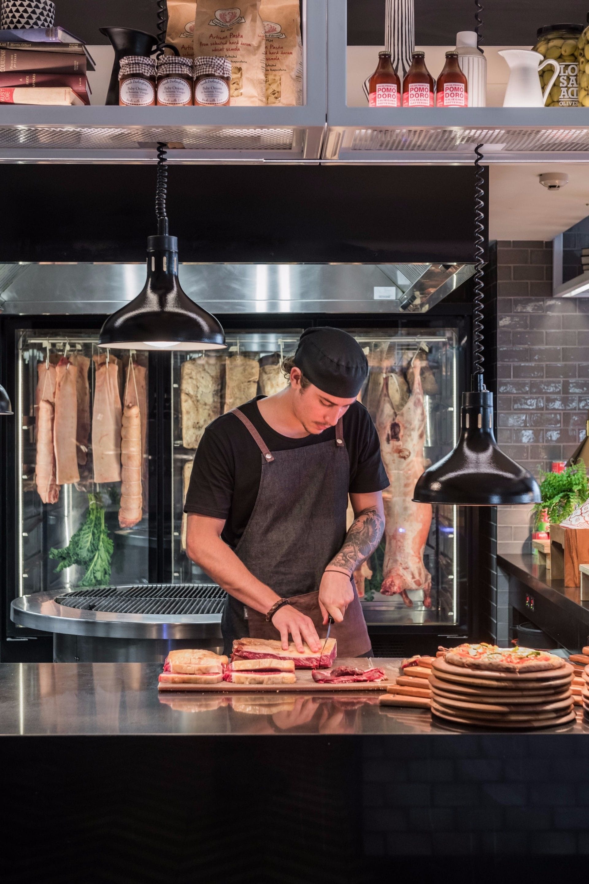 A chef preparing sandwiches in a kitchen with hanging meats and a pizza oven.