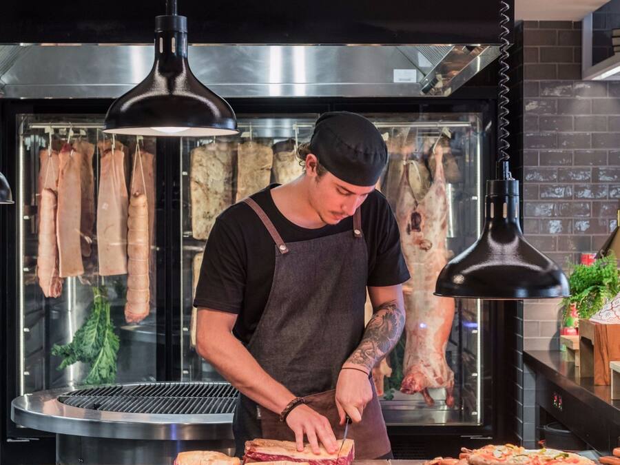 A chef preparing sandwiches in a kitchen with hanging meats and a pizza oven.