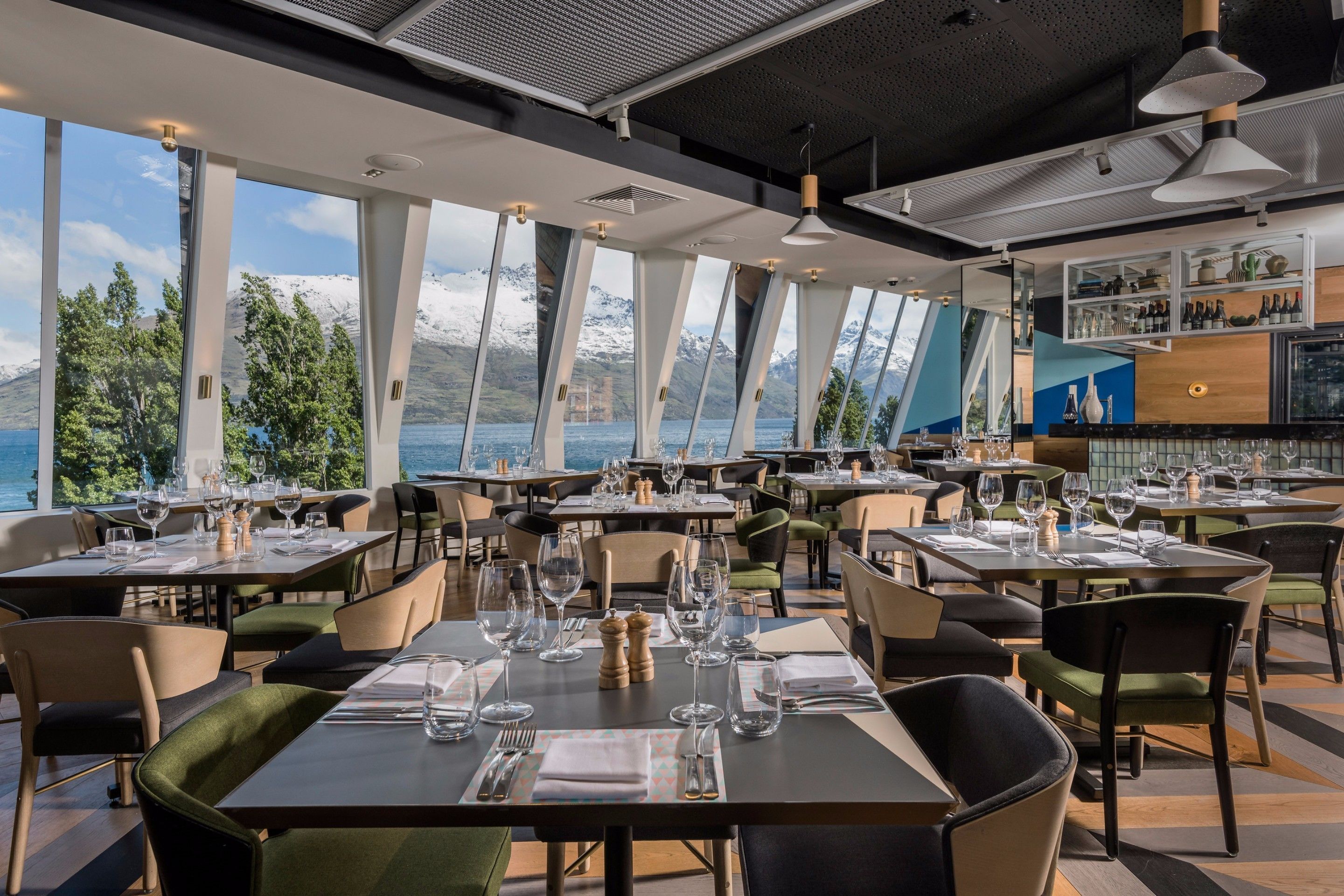A restaurant with tables set for dining, large windows offering a view of mountains and a lake, and a bar area with shelves of bottles.