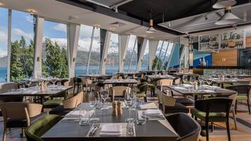 A restaurant with tables set for dining, large windows offering a view of mountains and a lake, and a bar area with shelves of bottles.