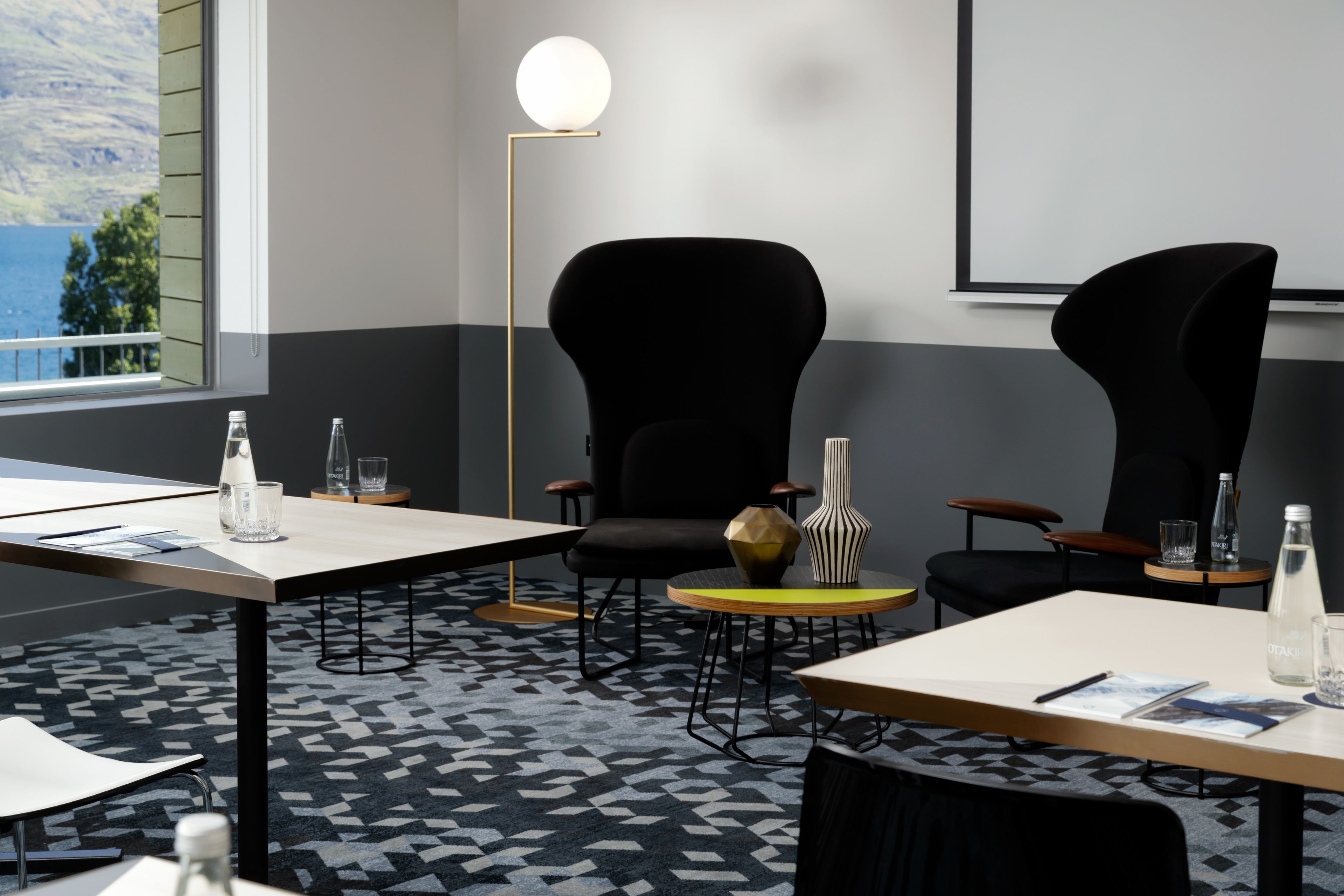 A conference room with two black chairs, a round table, a floor lamp, and a view of the outdoors.