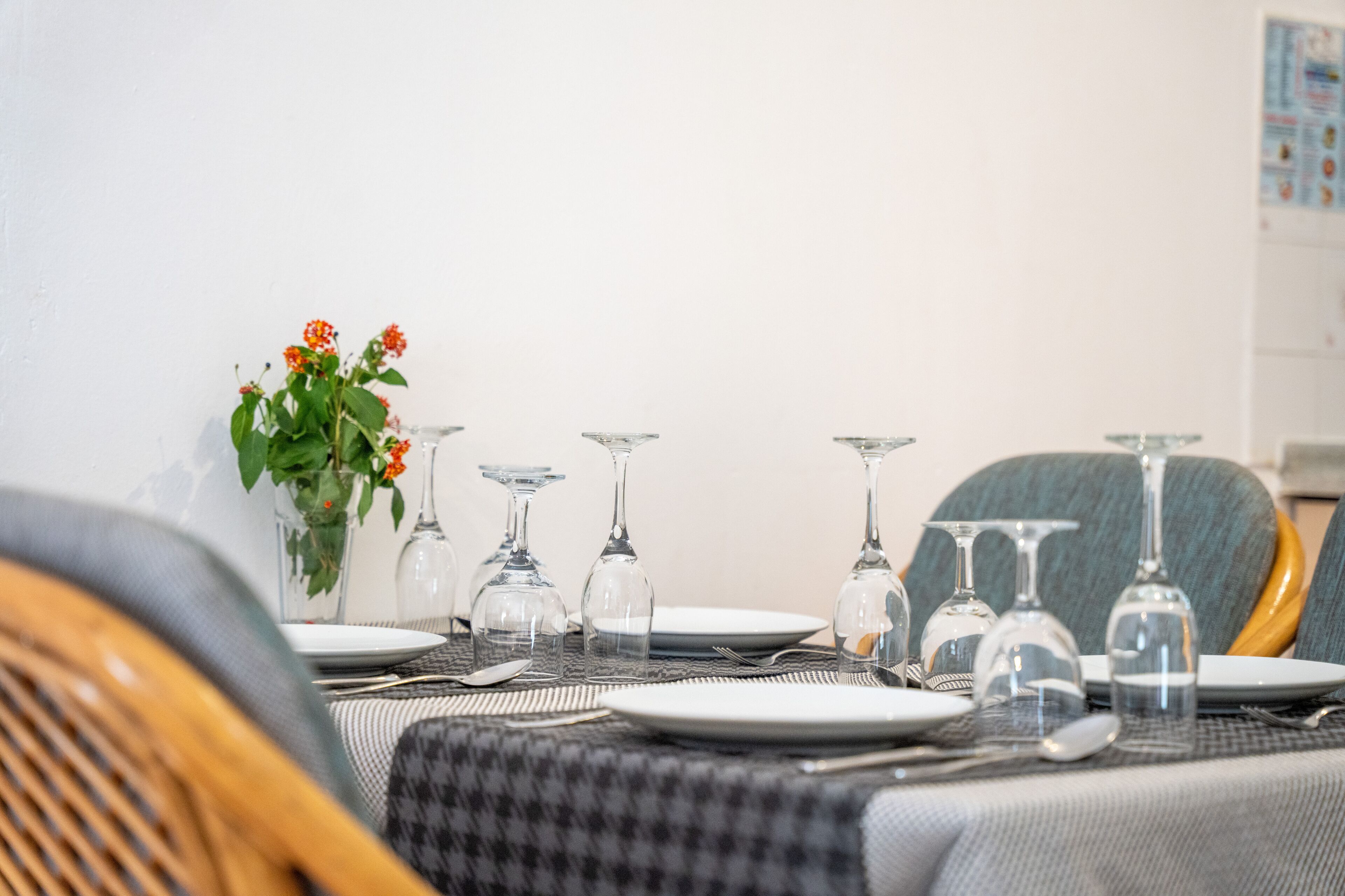 Una mesa de comedor dispuesta con platos, cubiertos y copas de vino, con un jarrón de flores a la izquierda.