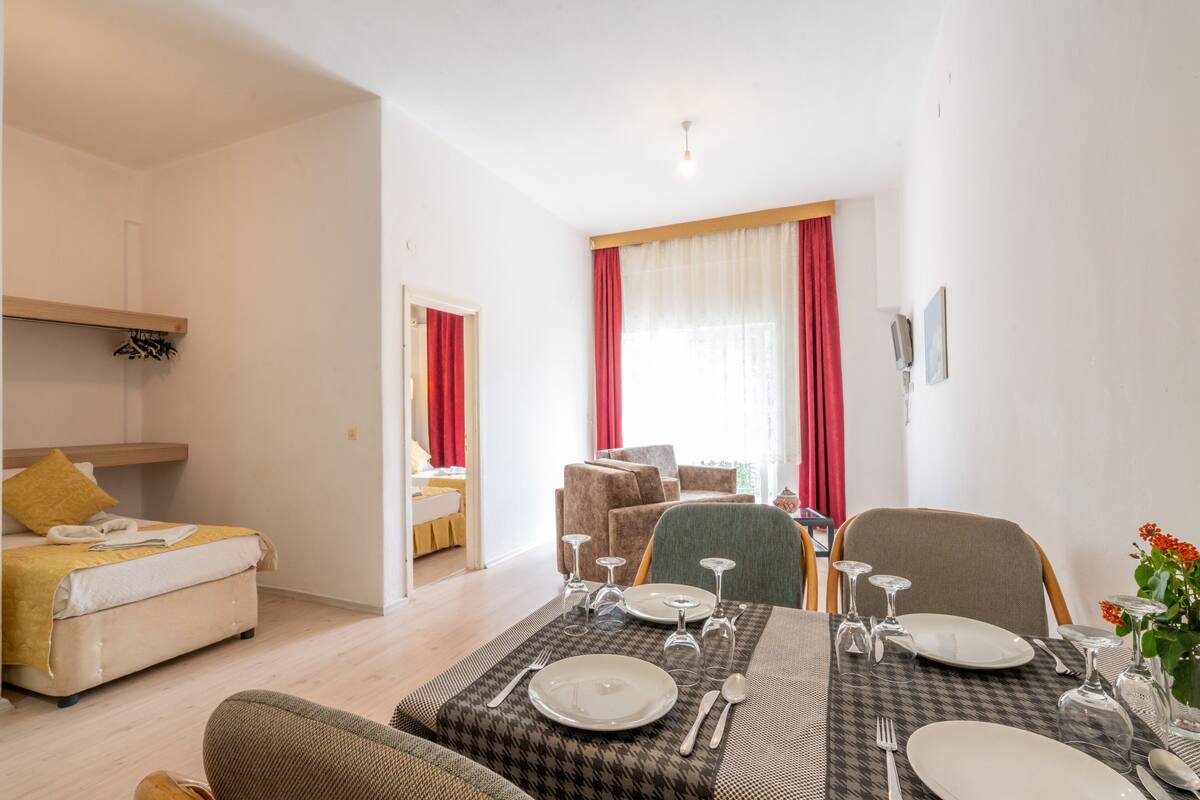 A well-lit room with a dining table set for four, a sofa, a bed, and a window with red curtains.