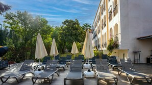 A sunlit outdoor lounge area with multiple lounge chairs and umbrellas, surrounded by lush greenery and a building with arched windows. - Grand Villa Sol Apart (Marmaris)