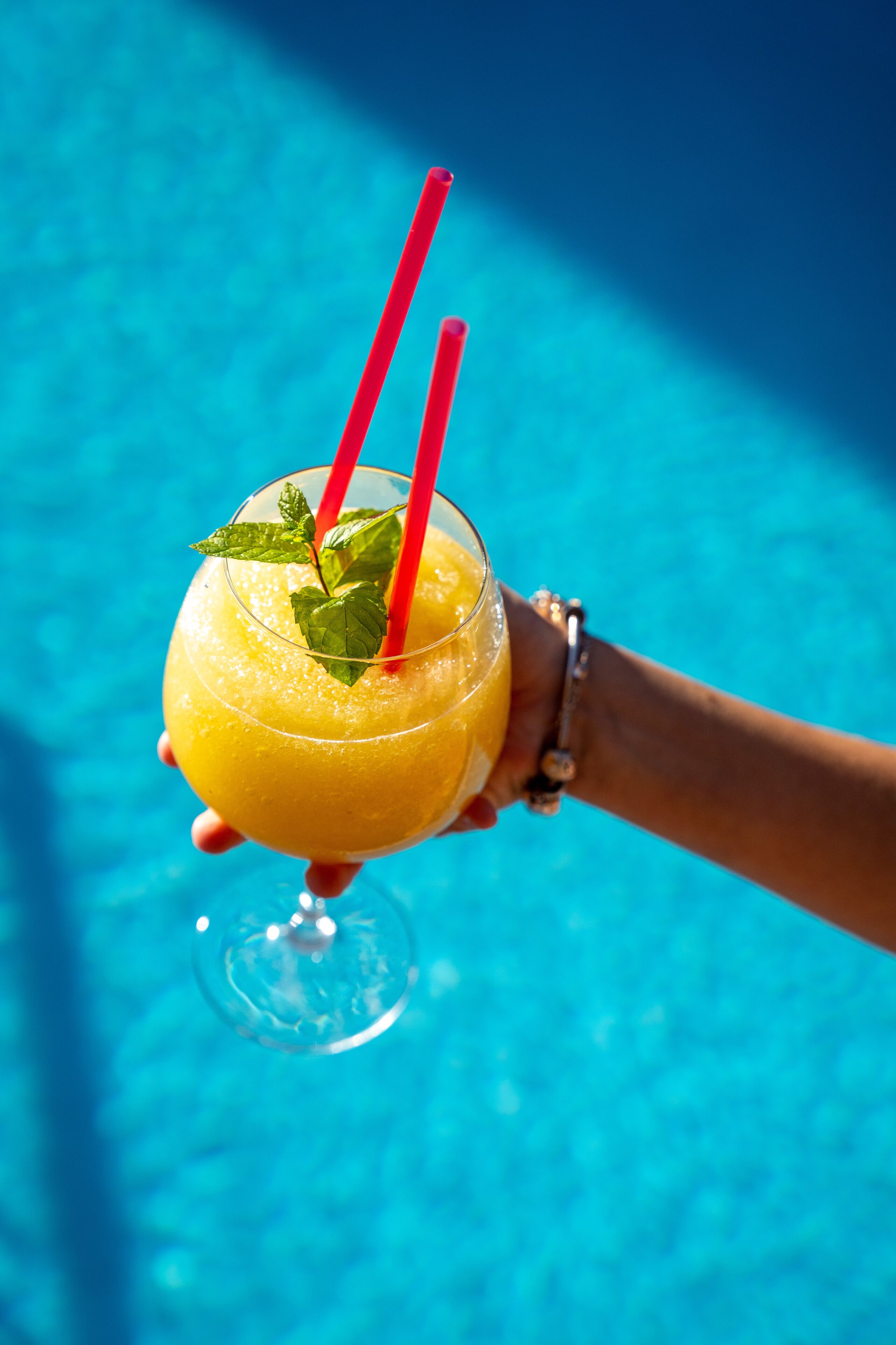 Eine Hand hält ein Glas mit einem gelben Getränk, zwei roten Strohhalmen und einem Minzblatt als Dekoration, vor einem blauen Pool im Hintergrund.